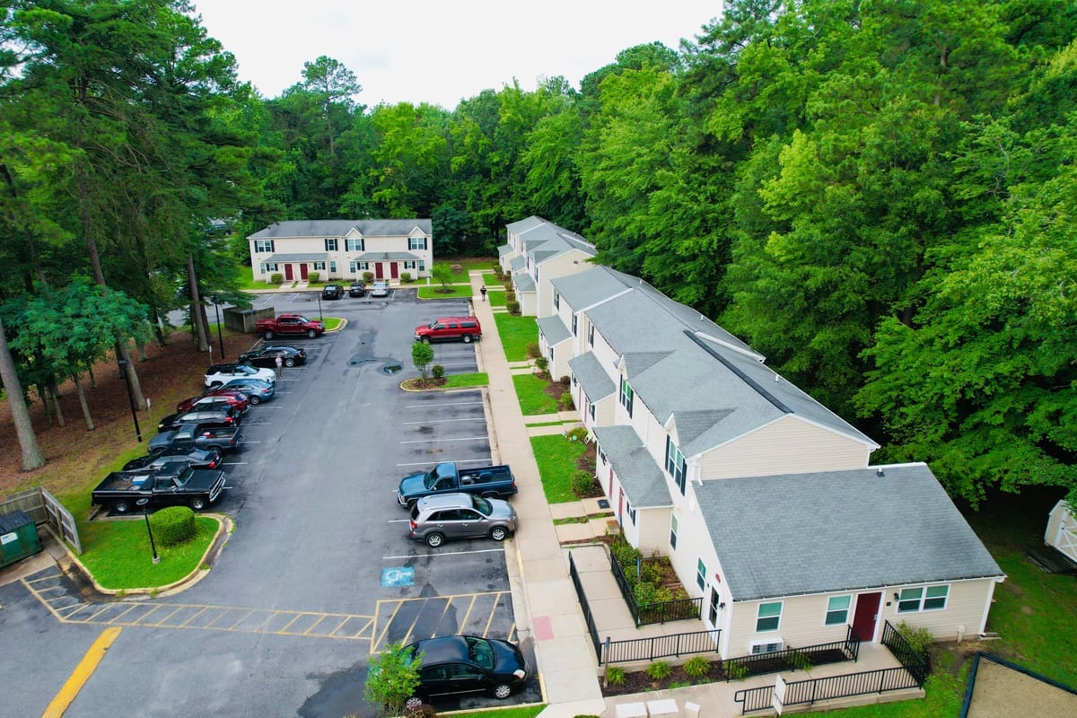 Aerial view of a row of two-story Colonial Beach Village townhomes with fenced patios along a parking lot, backed by trees