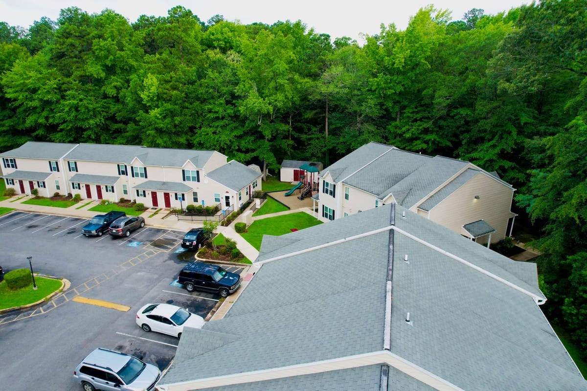 Aerial view of Colonial Beach Village showing several townhome buildings around a parking lot and a grassy courtyard with a playground, ringed by trees
